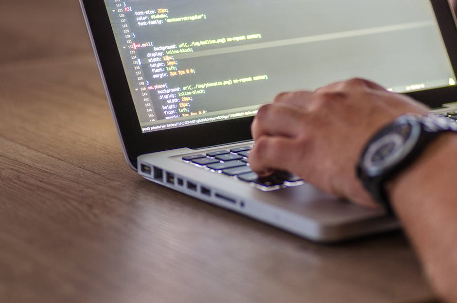 A close-up view of a person's hand typing on a silver laptop keyboard placed on a wooden surface, displaying lines of code with a dark background and colorful syntax highlighting. The person is wearin