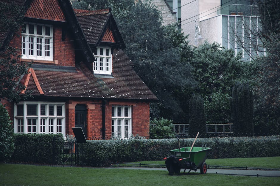 A red brick house with a steeply pitched tiled roof and white-framed windows surrounded by a well-maintained garden. The garden features a neatly trimmed hedge lining a paved pathway, with lush green